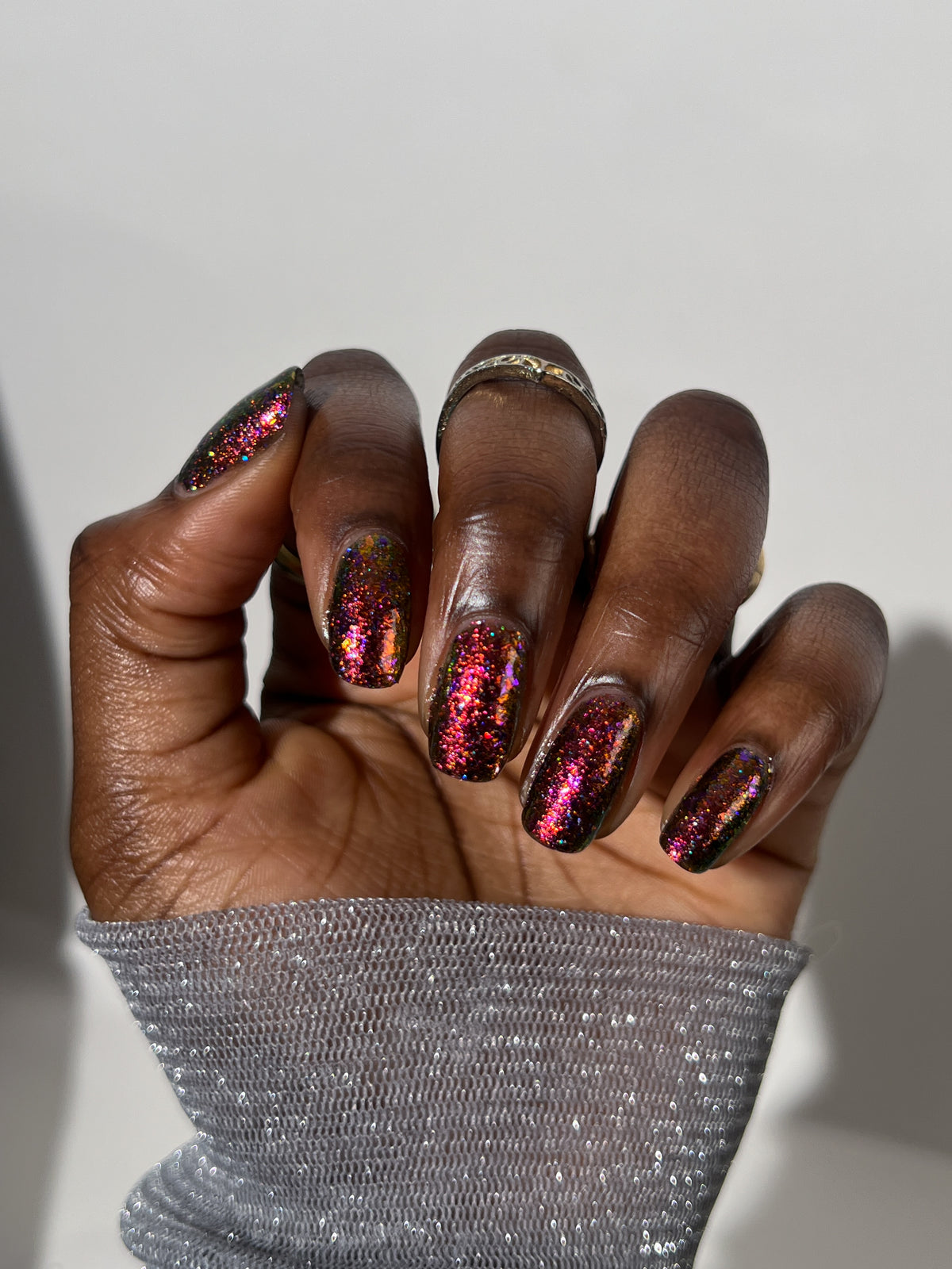 A close-up of a hand with manicured nails painted in a sparkling holographic pink and gold glitter polish, wearing a silver glittery sleeve against a plain light gray background.