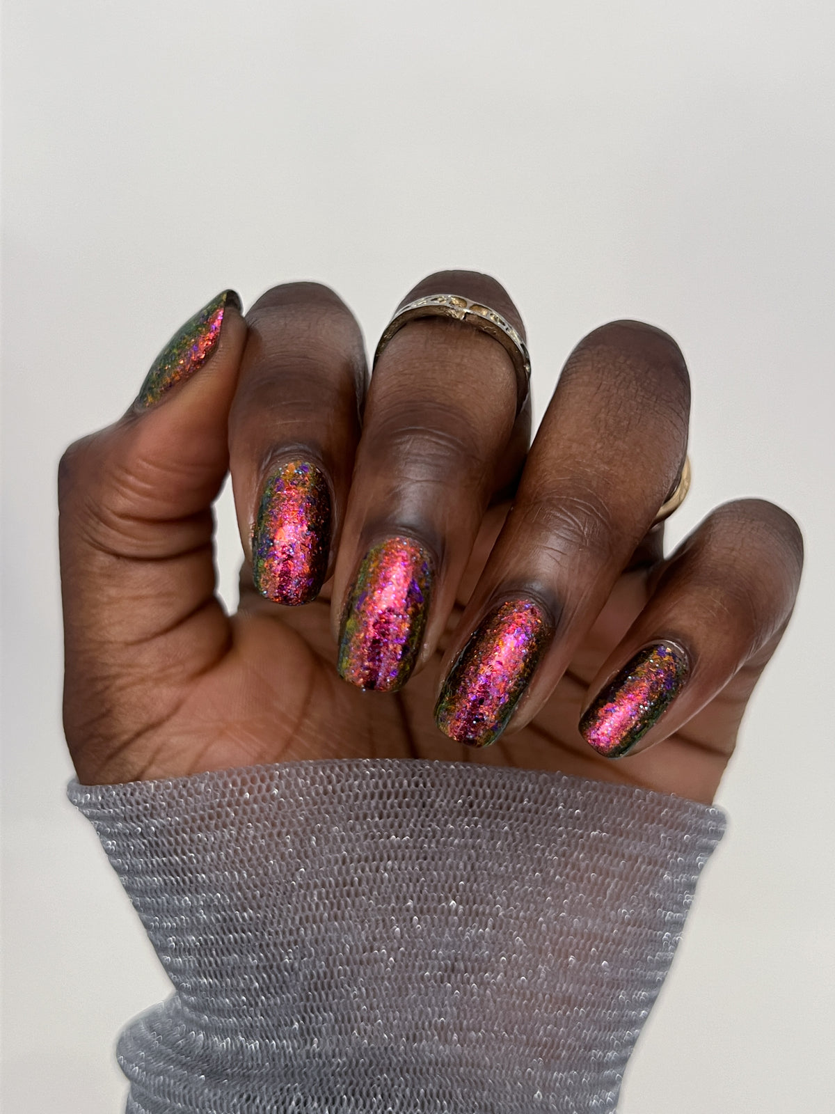 A close-up of a hand with long, glossy almond-shaped nails painted in a shimmering holographic pink, purple, and green glitter nail polish, wearing a silver knit cuff and gold rings against a plain light gray background.