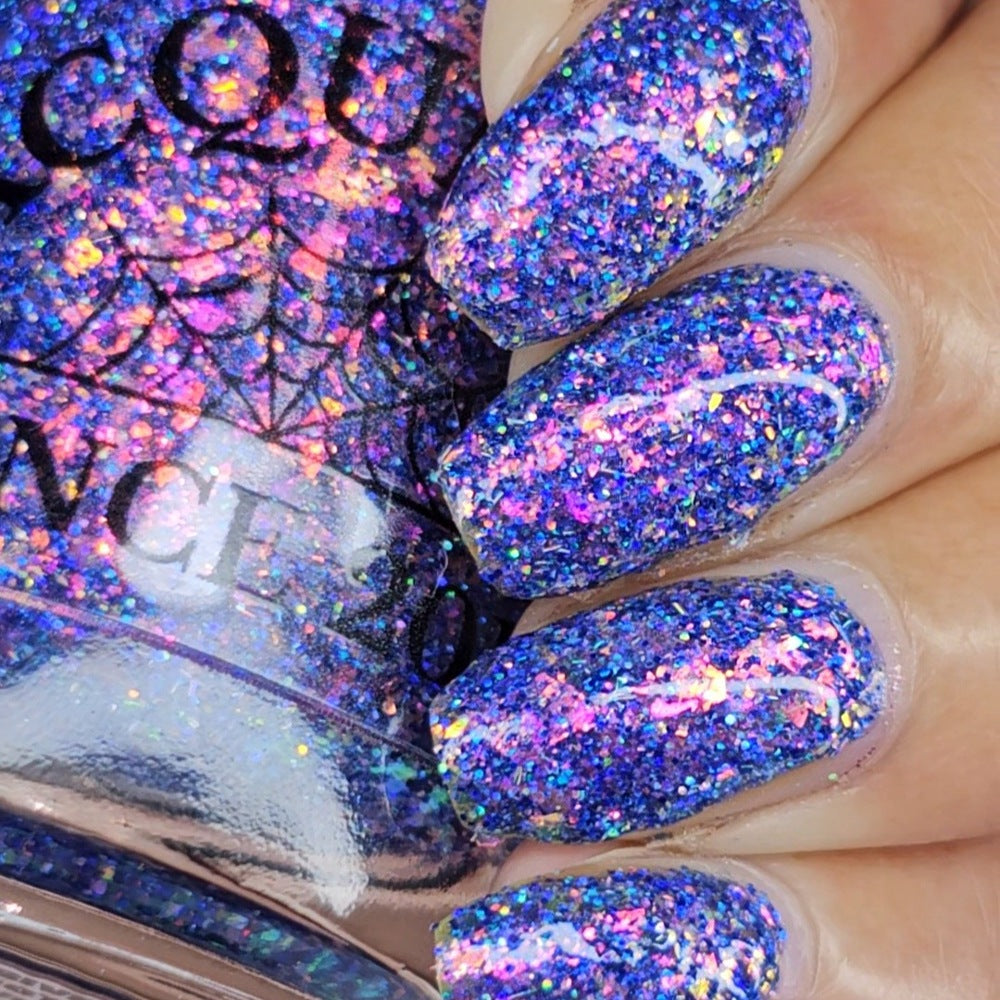 Close-up of a hand with fingernails painted in a dense holographic glitter polish that shifts between blue, purple, pink, and gold, next to a bottle of the same nail polish.