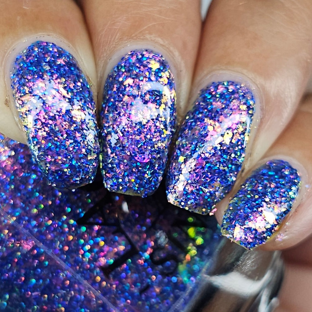 Close-up of a hand with manicured nails painted in a dense, iridescent holographic glitter nail polish showing shades of blue, purple, and pink, with a bottle of the glitter polish visible in the foreground.