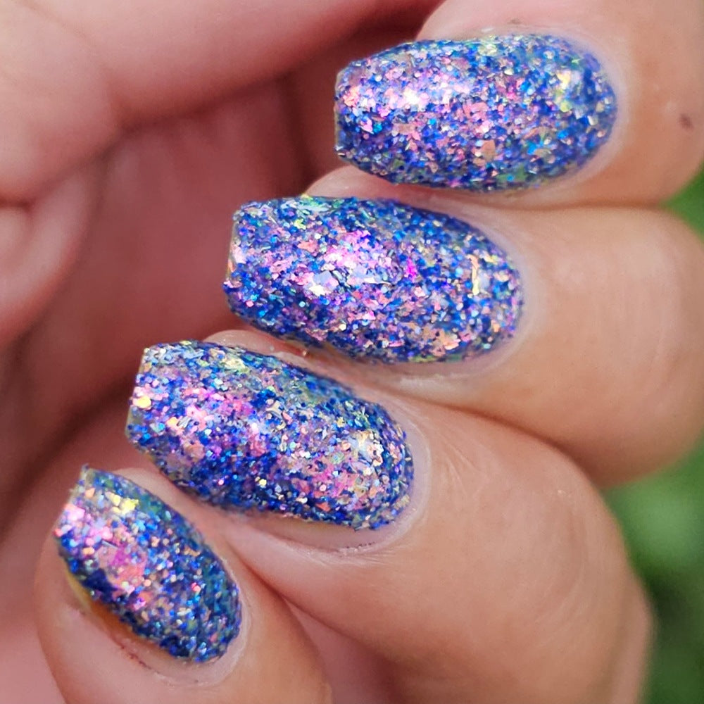 Close-up of a hand with manicured nails painted in a glittery blue polish with holographic pink, purple, and iridescent flecks.