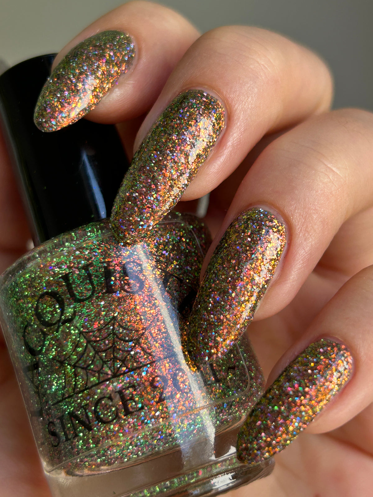 nails painted with sparkly glittery polish showing pink, red, green, gold and orange glitters in a darkened base color with green reflective glitter. holding lacquistry bottle