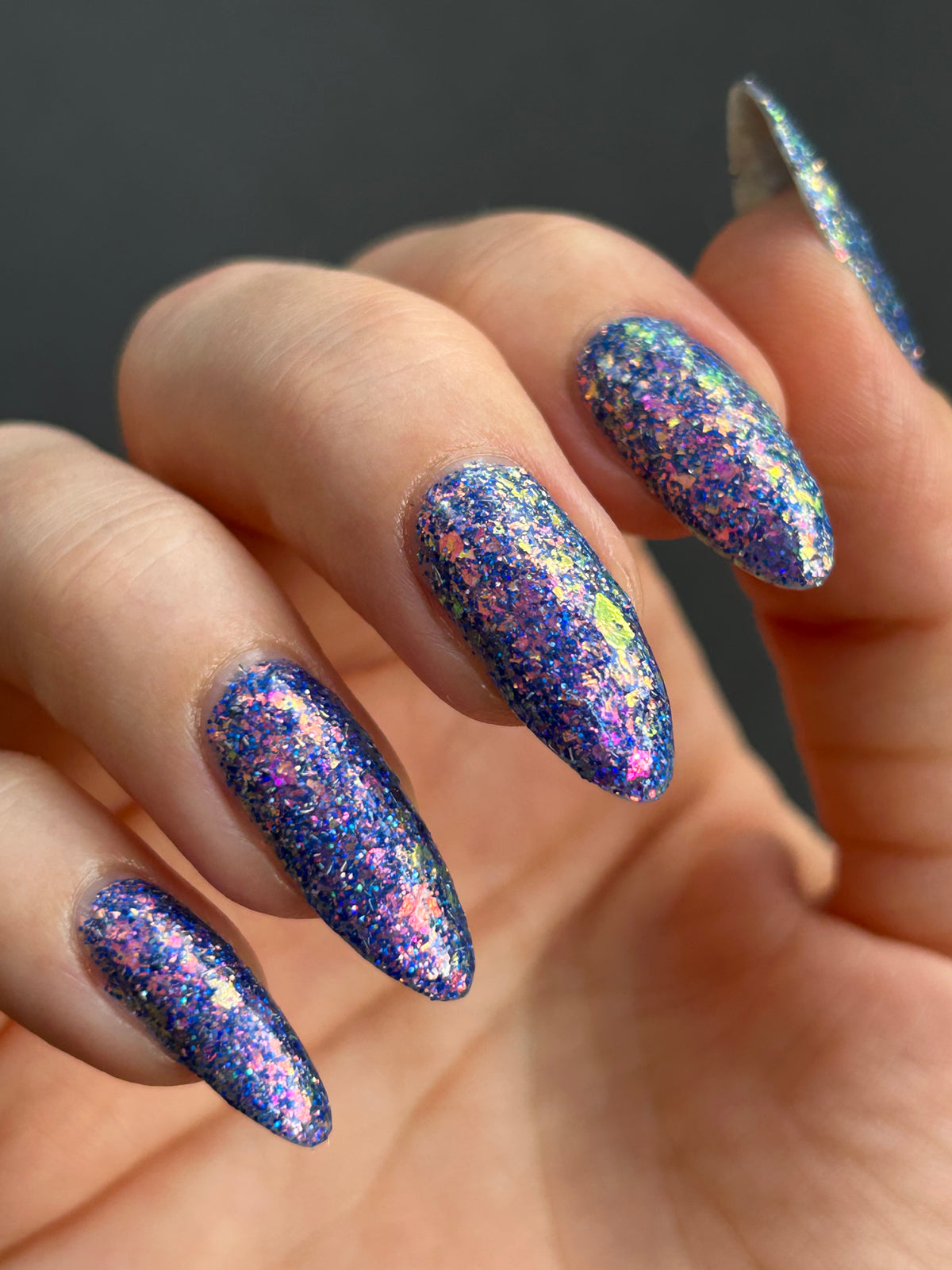 A close-up of a hand with long, almond-shaped nails painted in a sparkling holographic glitter nail polish that shifts between blue, purple, pink, and gold against a dark gray background.