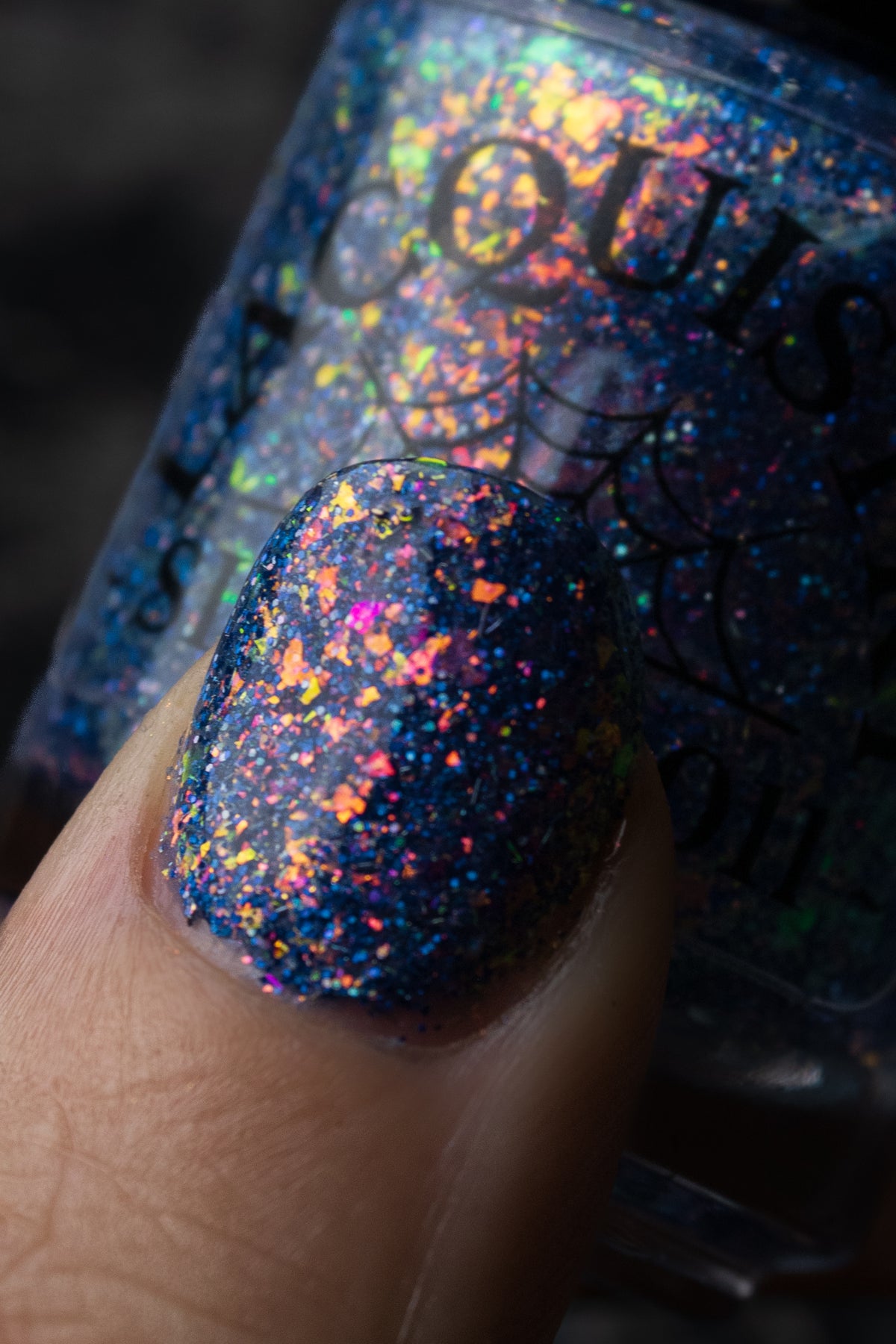 A close-up of a fingernail painted with a dark navy glitter nail polish featuring holographic rainbow flecks, with a bottle of the same polish blurred in the background.