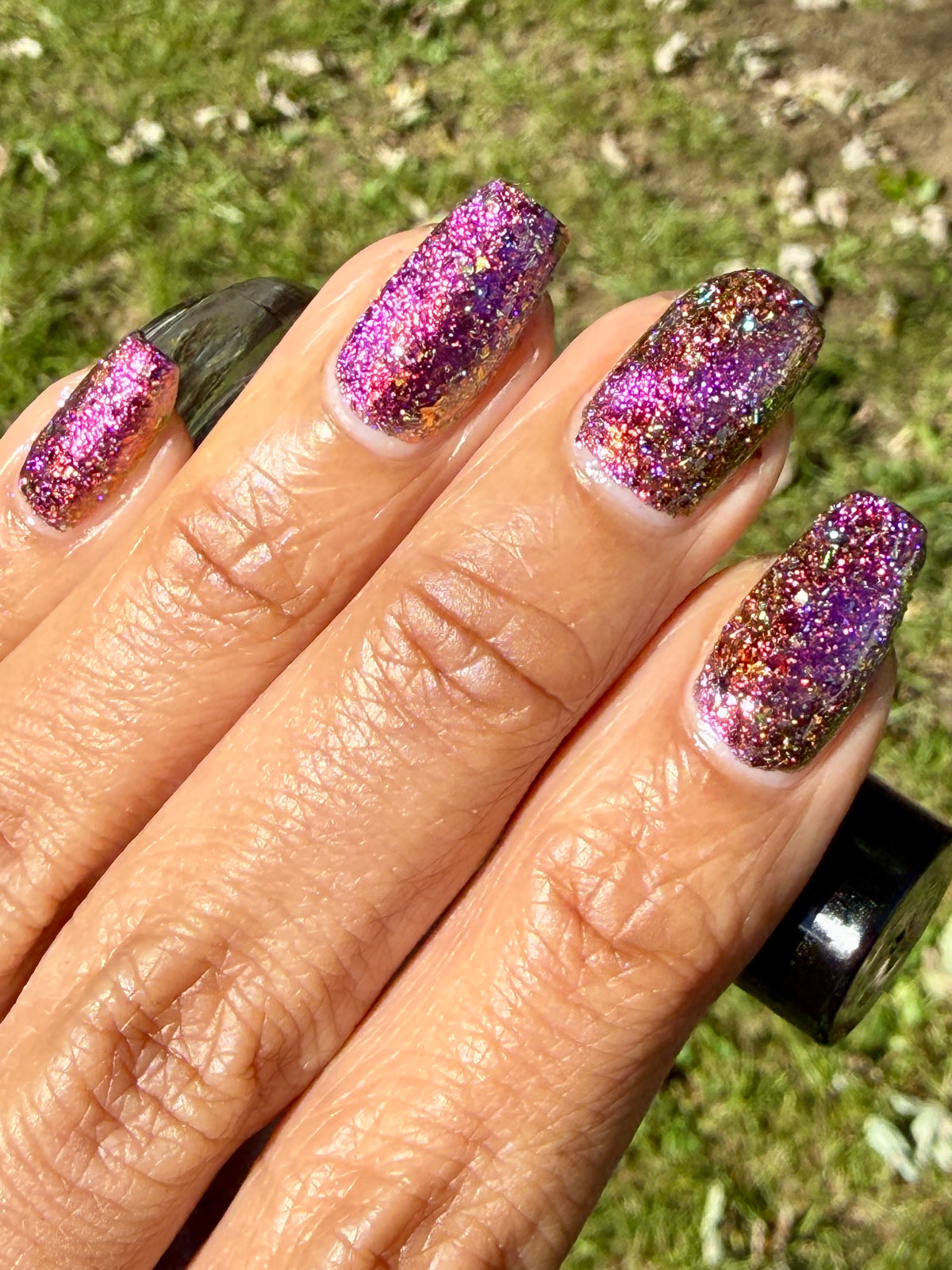 flakie magnetic polish showing red, orange, gold, green flakes, holo glitter, and magnetic effect of fuchsia to purple with grass background