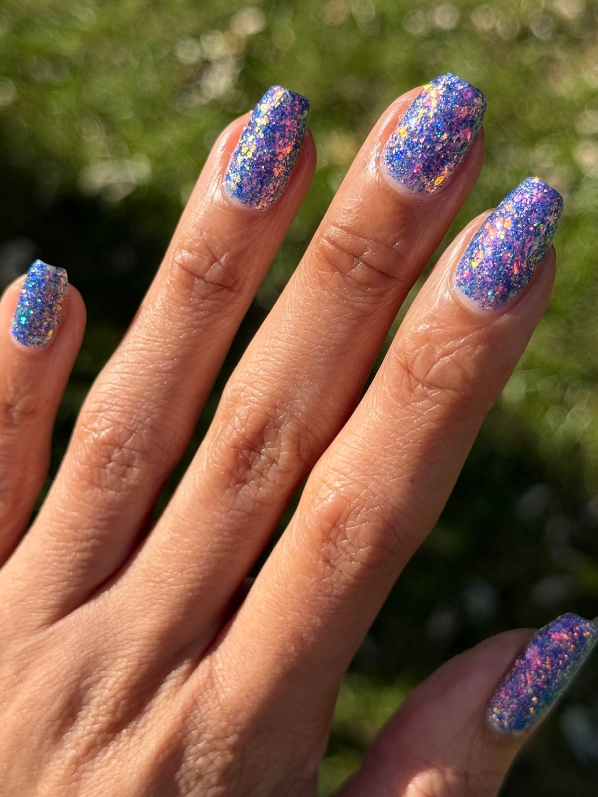 A close-up of a hand with manicured nails painted in a glittery blue and purple holographic nail polish, set against a blurred green outdoor background.