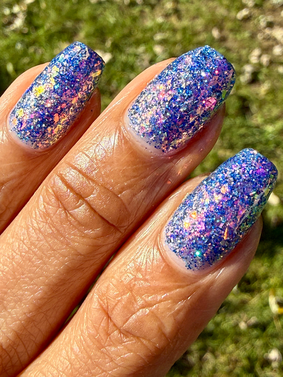 A close-up of a hand with long, almond-shaped nails painted in a glittery blue nail polish that reflects purple, pink, and gold tones, set against a blurred green grass background.
