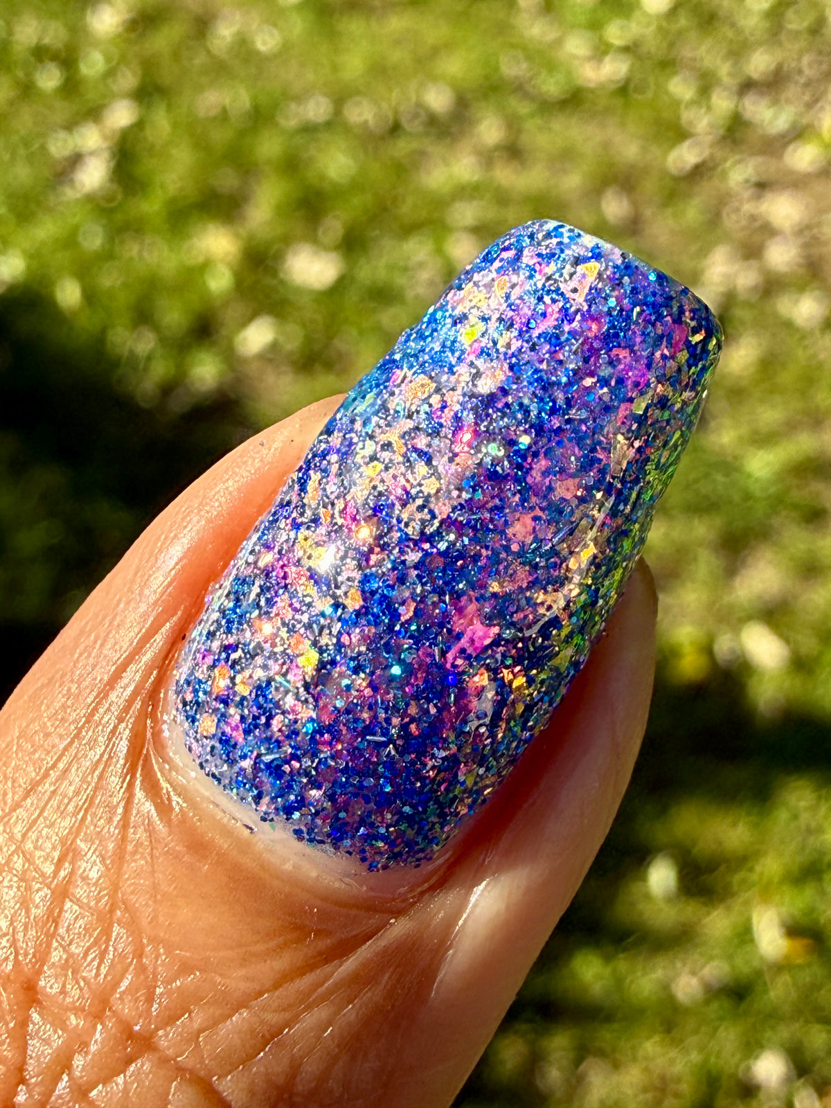 A close-up of a fingernail painted with a dense, glittery holographic nail polish that shifts between blue, purple, pink, and green, set against a blurred green grassy background.