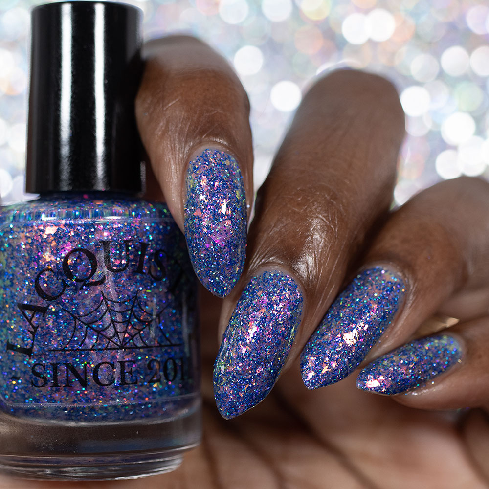 A close-up of a hand with long, almond-shaped nails painted in a sparkling holographic blue nail polish, holding a bottle of the same glittery blue polish. The background is a soft, out-of-focus bokeh of silver and white lights.