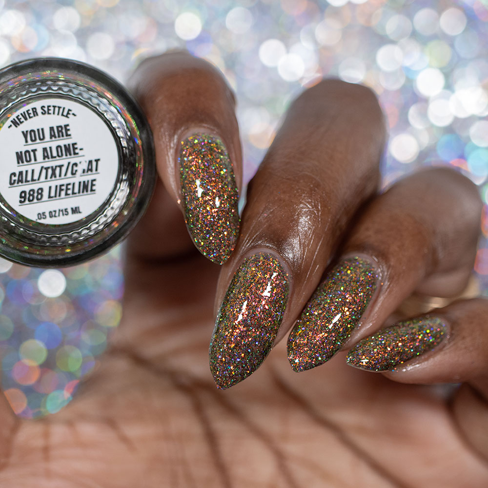 nails painted with sparkly glittery polish showing pink, red, green, gold and orange glitters in a darkened base color with green reflective glitter. bottle says you are not alone 988 lifeline