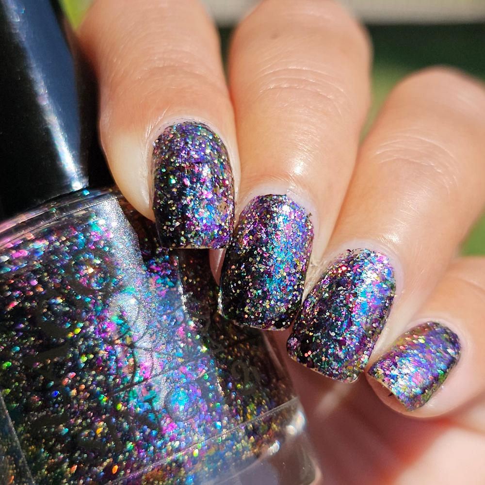 A close-up of a hand with nails painted in a dark holographic glitter nail polish, holding a matching nail polish bottle. The polish reflects rainbow iridescent glitter.