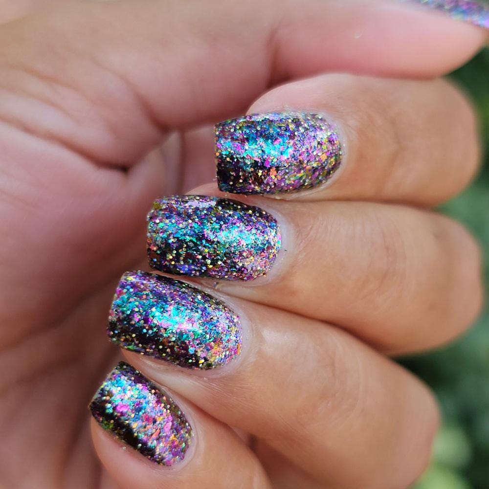 A close-up of a hand with nails painted in a dark holographic glitter nail polish, holding a matching nail polish bottle. The polish reflects rainbow iridescent glitter.