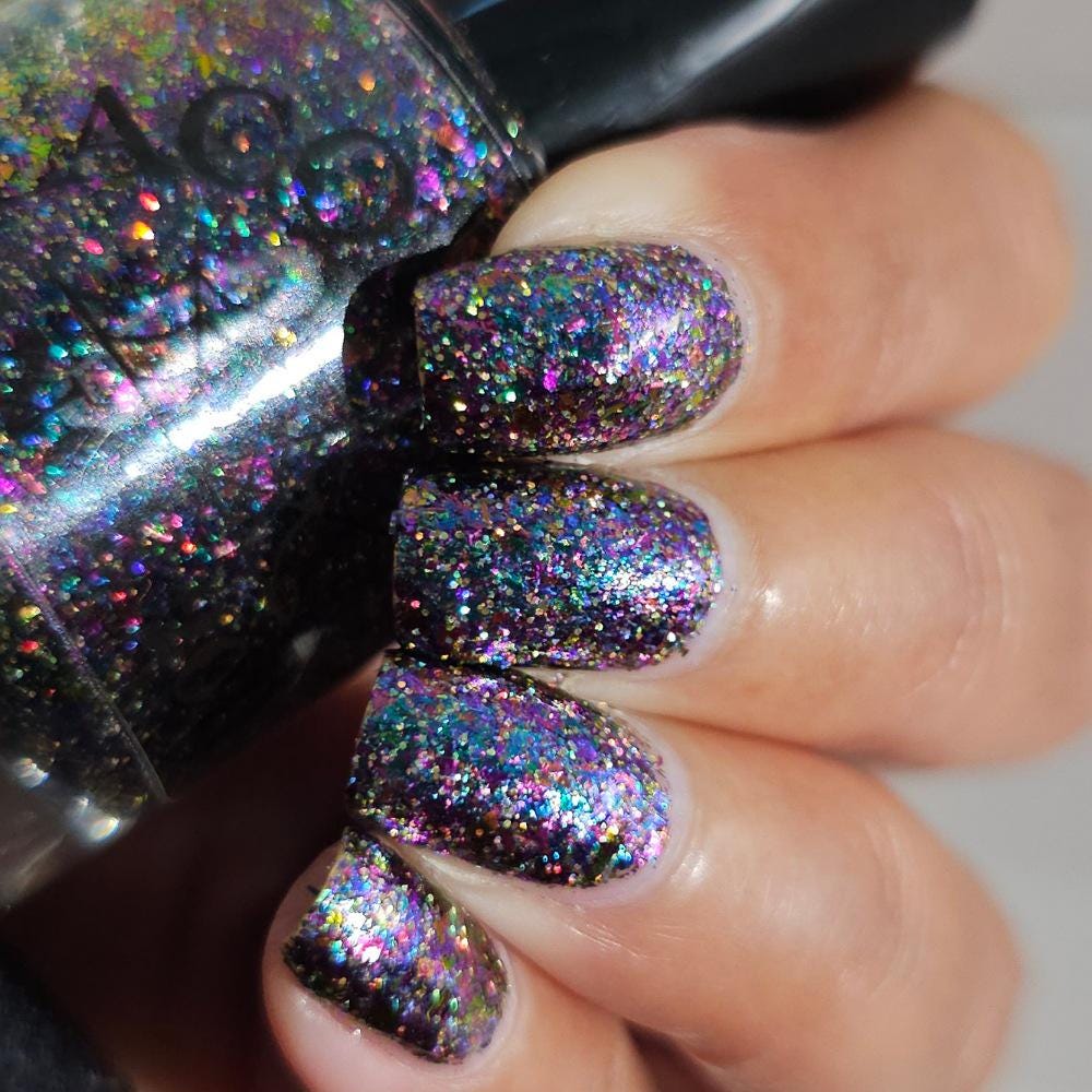 A close-up of a hand with nails painted in a dense, iridescent holographic glitter polish, with the matching glitter nail polish bottle shown in the foreground.