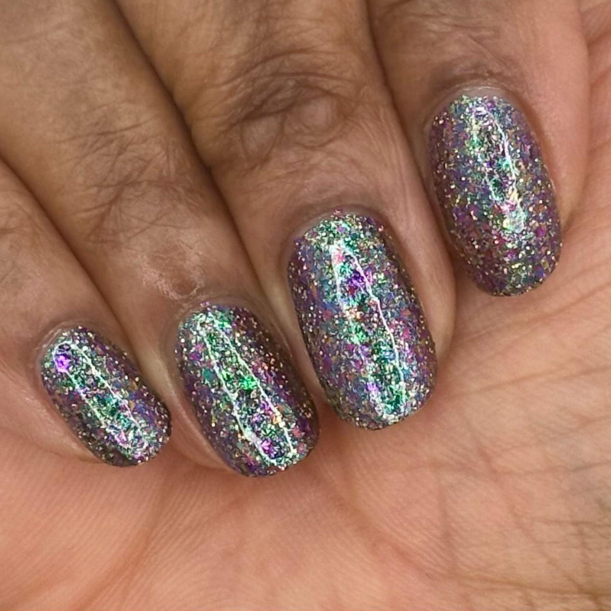 A close-up of a hand with nails painted in a dark holographic glitter nail polish, holding a matching nail polish bottle. The polish reflects rainbow iridescent glitter.