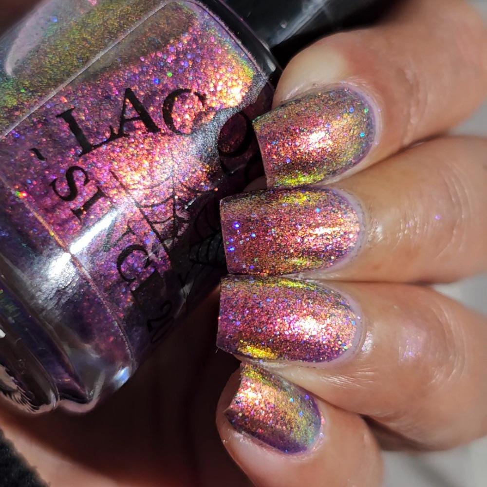 medium skin toned hand with medium/long square nails wearing metallic purple pink sparkle nail polish holding matching bottle of light purple nail polish