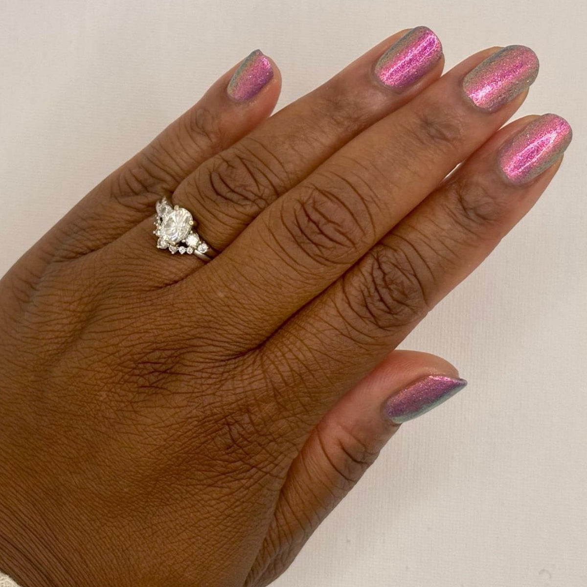 A close-up of a hand with a darker skin tone with purple pink holographic nail polish and a silver floral ring, shown against a plain off-white background.
