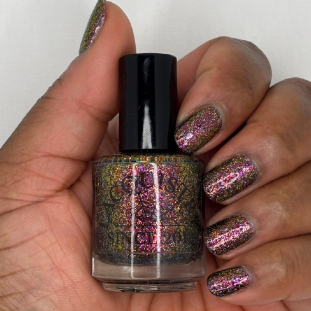 A hand with a dark skin tone holding a bottle of nail polish with matching glittery pink, burgundy, green, and gold holographic nail polish applied, against a plain light background