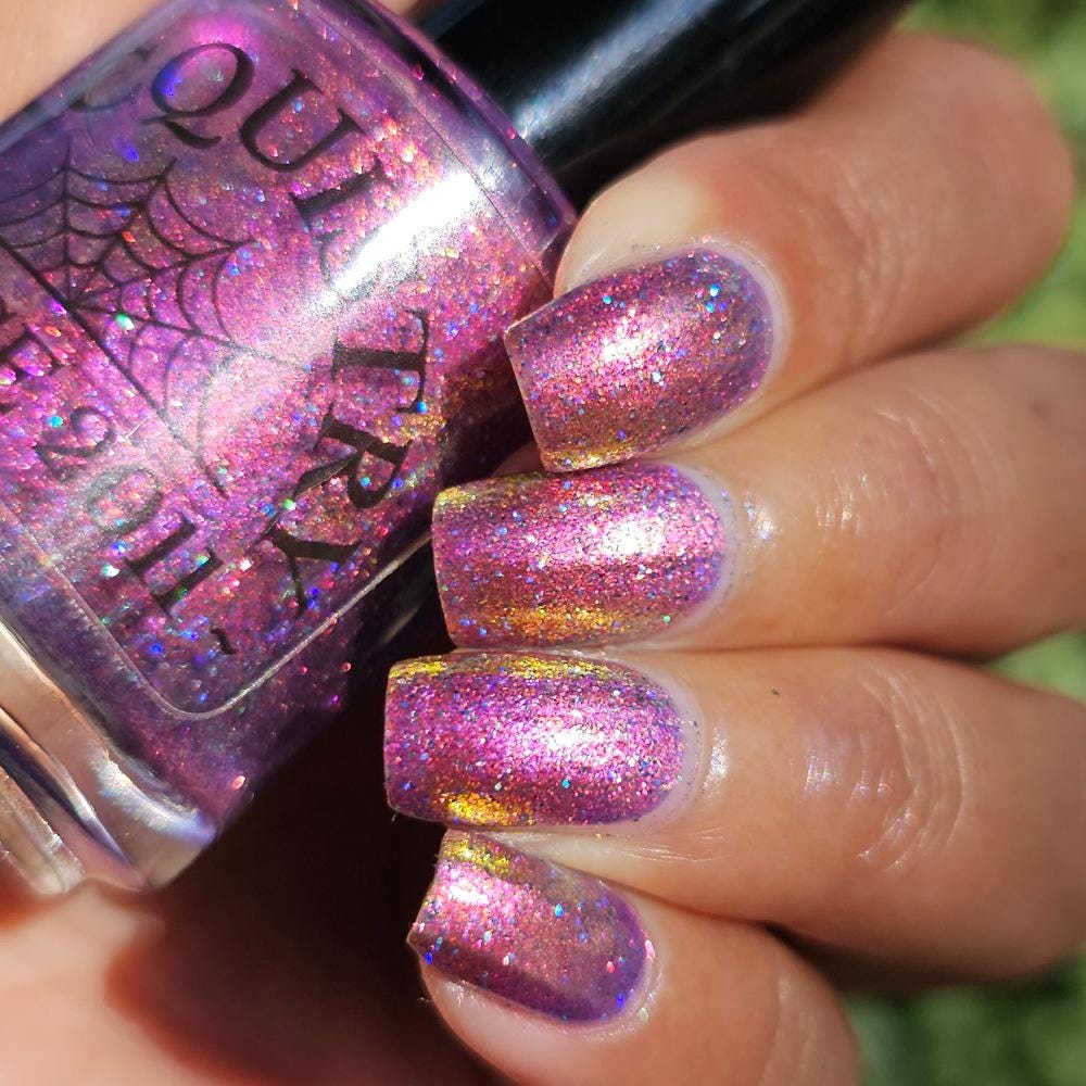 medium skin toned hand with medium/long square nails wearing metallic purple pink sparkle nail polish holding matching bottle of light purple nail polish