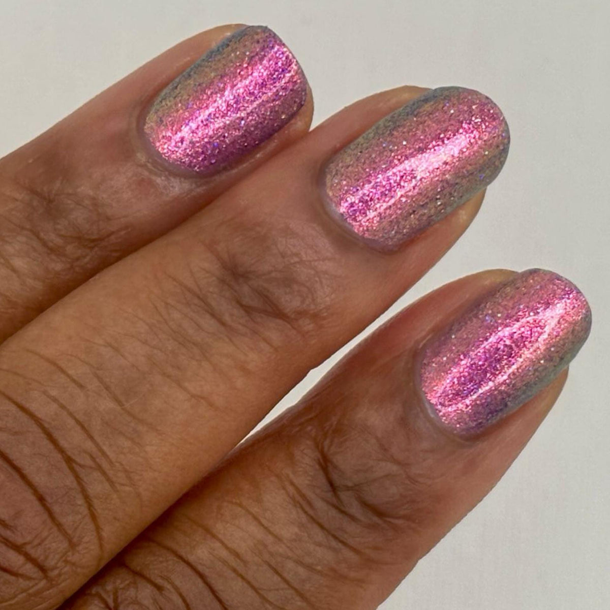 darker skin toned hand with short nails wearing metallic purple pink sparkle nail polish 