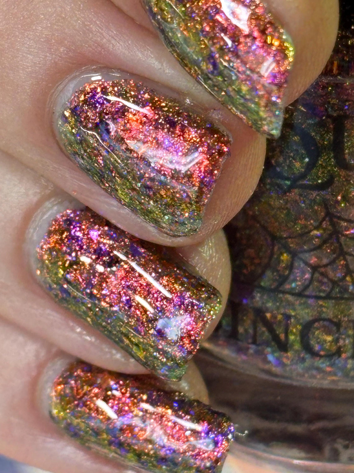 Close-up of a hand with nails painted in a chunky, glittery holographic nail polish showing iridescent pink, purple, green, and gold shimmer, with part of the bottle label visible in the background.