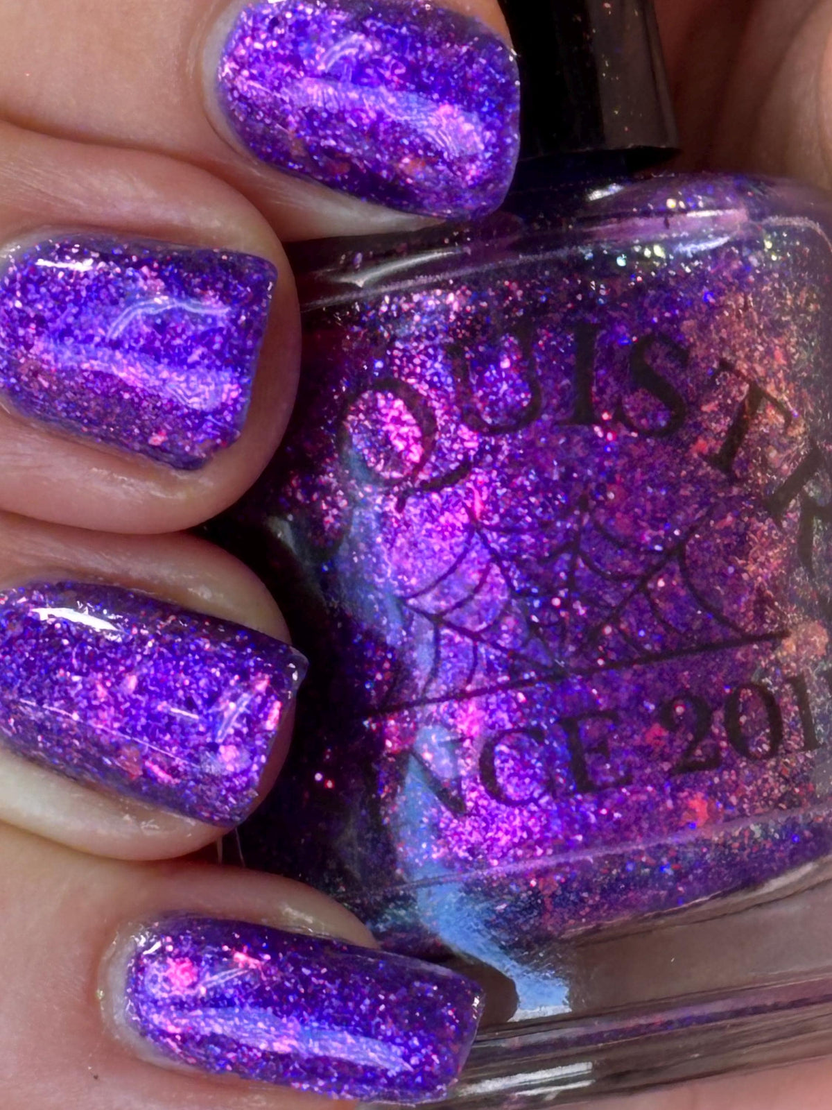 A close-up of a hand with fingernails painted in a glittery purple nail polish, holding a matching purple glitter nail polish bottle. The bottle label shows the brand name 'Lacquistry'. light skin.
