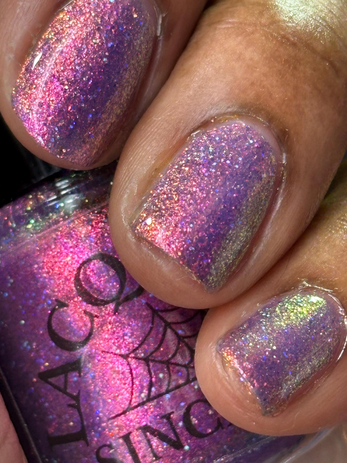 darker skin toned hand with short nails wearing metallic purple pink sparkle nail polish holding matching bottle of light purple nail polish