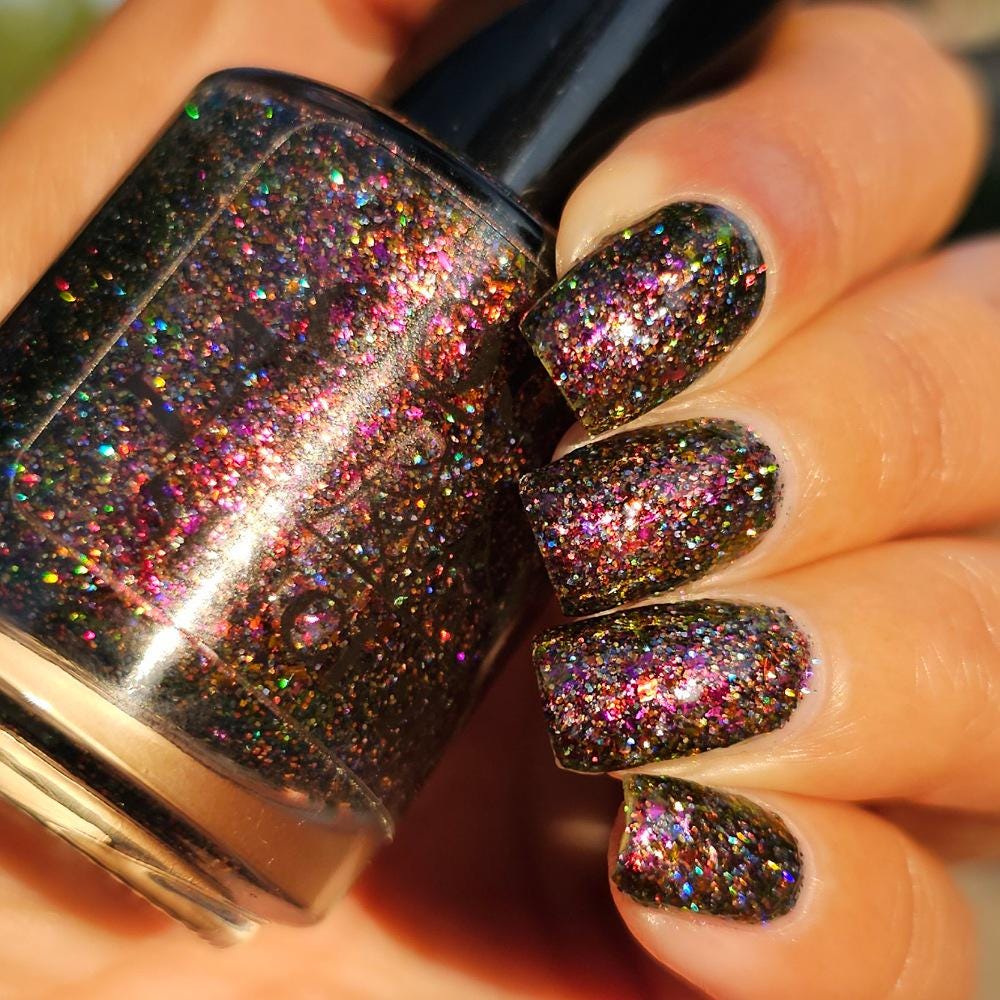 A close-up of a medium skin tone hand with manicured nails painted in a dark multichrome burgundy glitter nail polish that reflects holographic rainbow sparkle, holding a matching glitter nail bottle in the sun