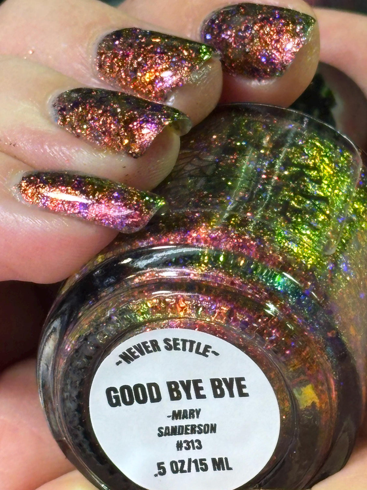 A close-up of a hand with glittery iridescent nail polish applied, holding a bottle labeled 'Good Bye Bye' by Mary Sanderson. The polish reflects holographic rainbow shimmer over a dark base.
