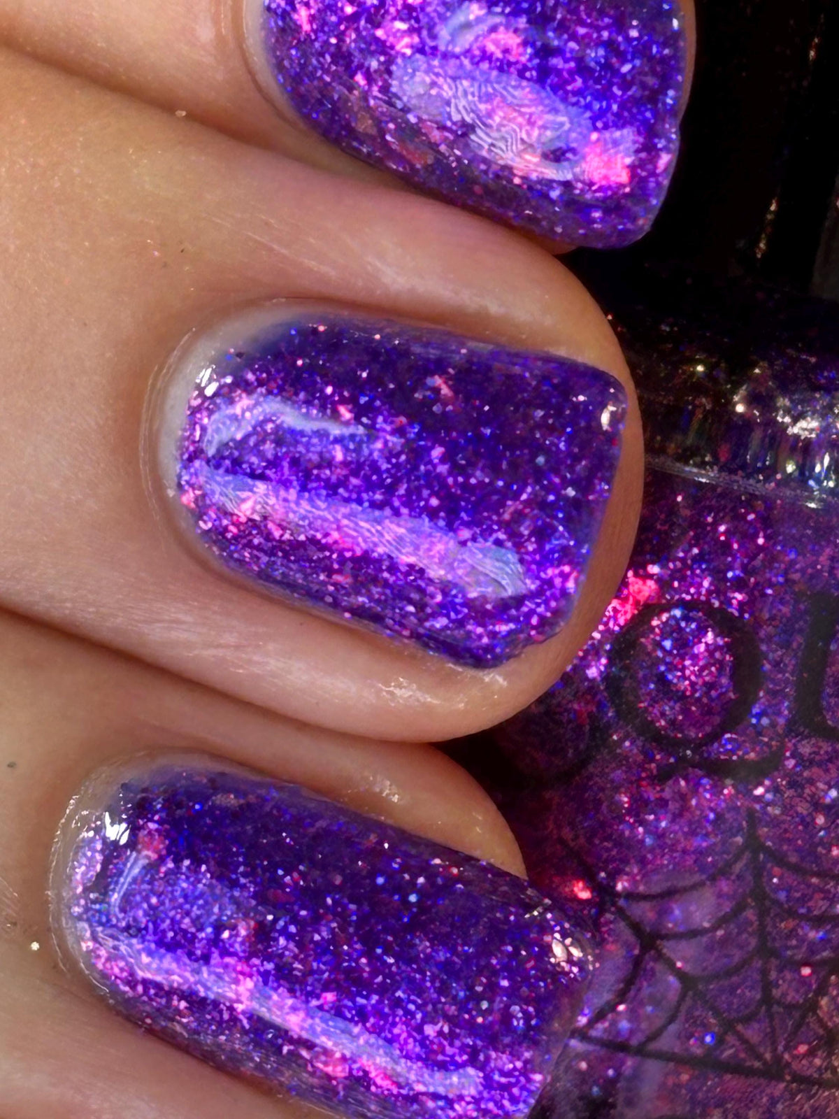 Close-up of fingernails painted with a glittery purple and pink holographic nail polish, with part of the matching bottle visible in the background.