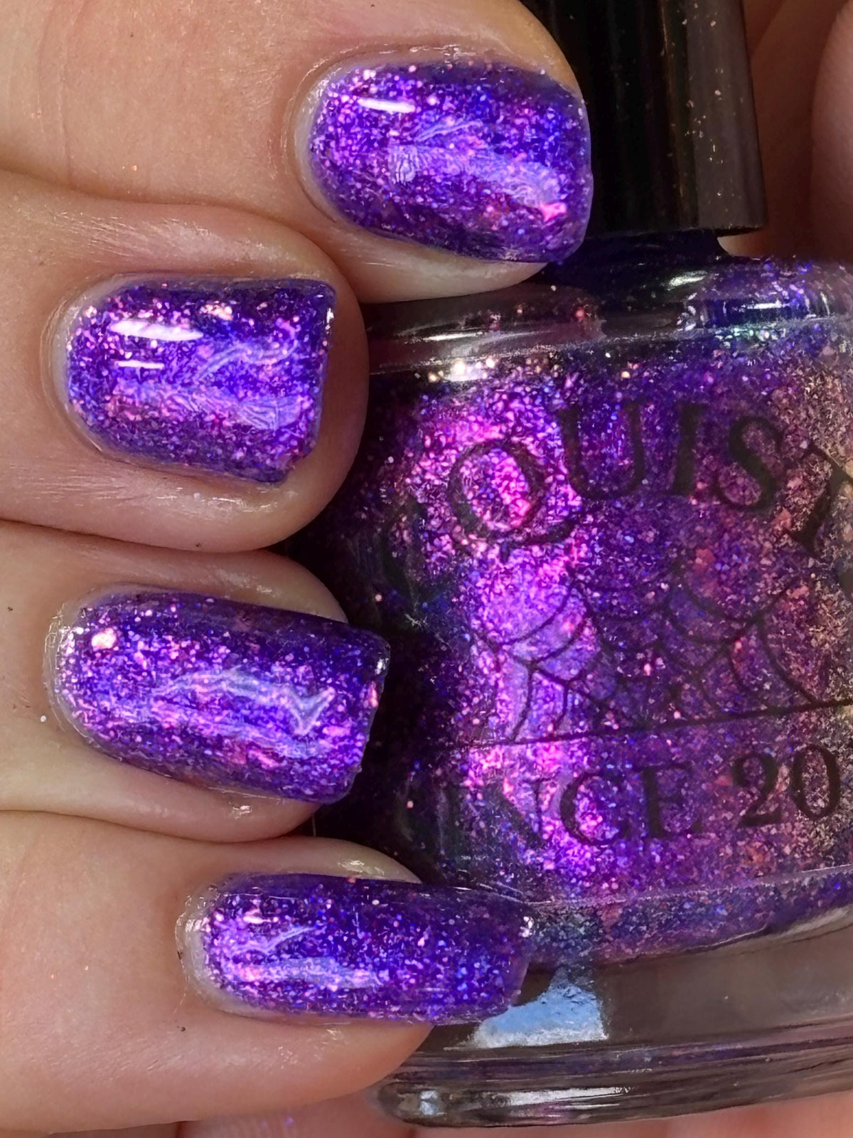 closeup of nails painted vibrant purple and violet sparkly nail polish holding a bottle of the same shade with the words "lacquistry since 2011" printed on the bottle, light skin tone