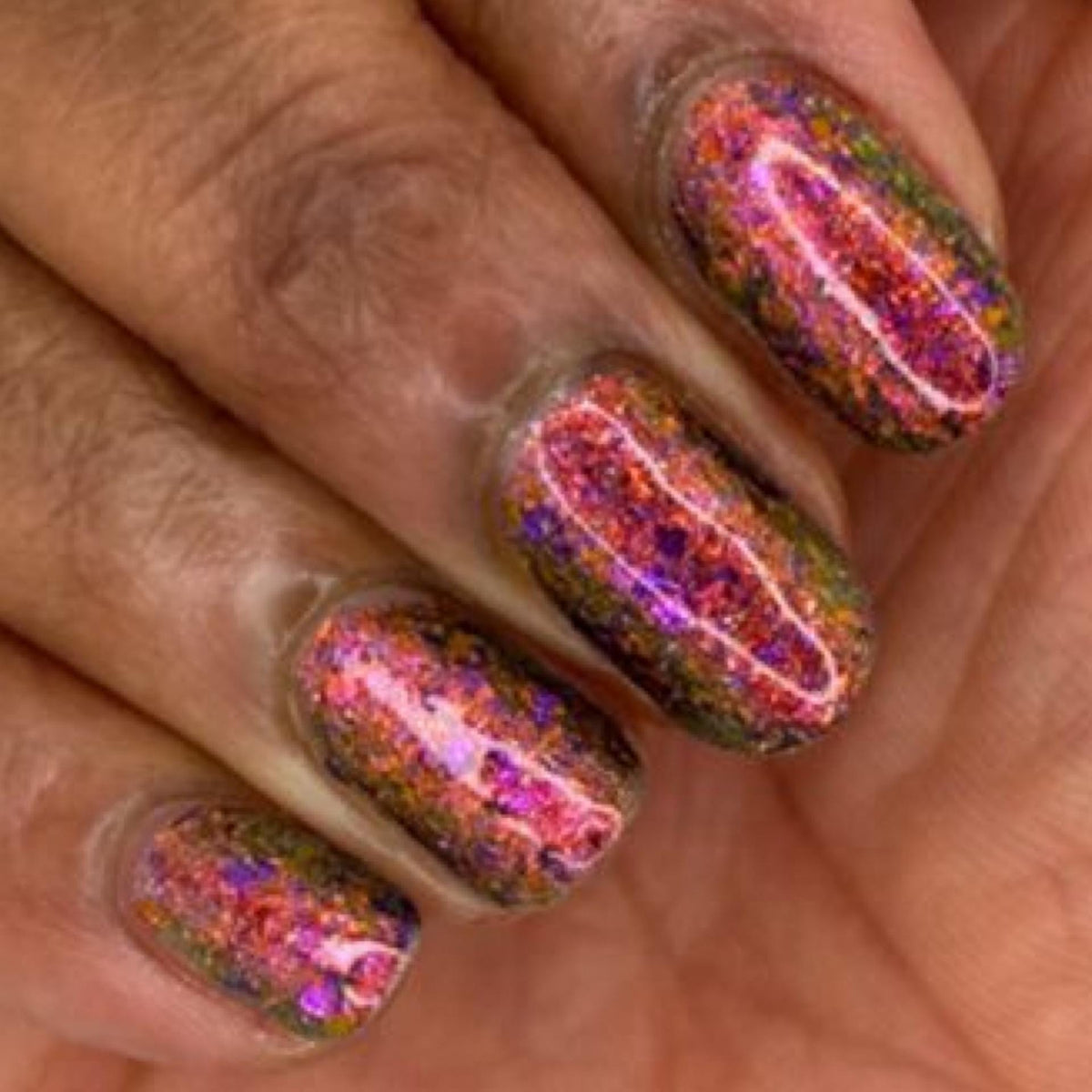 Close-up of a hand with manicured nails painted in a holographic glitter nail polish showing pink, purple, orange, and green iridescent reflections.