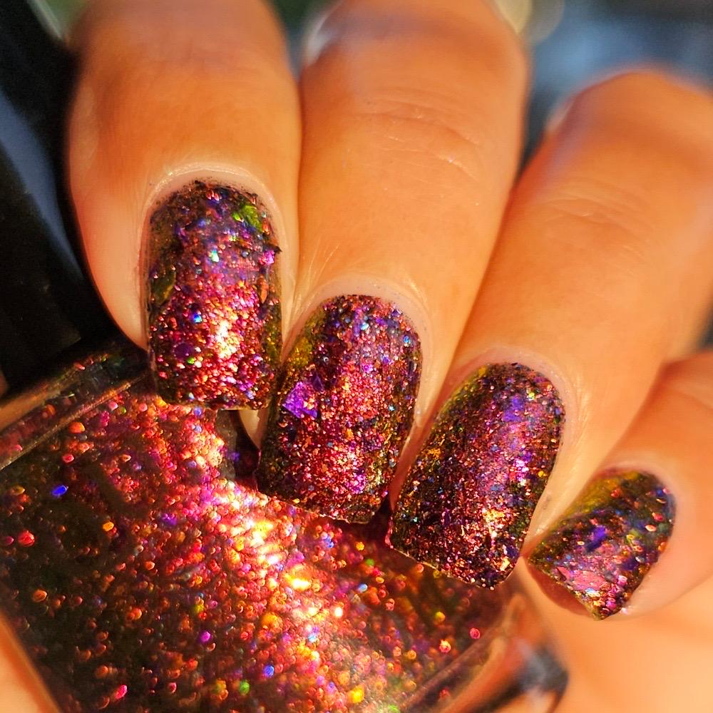 Close-up of a hand with manicured nails painted in a holographic glitter nail polish showing pink, purple, orange, and green iridescent reflections.