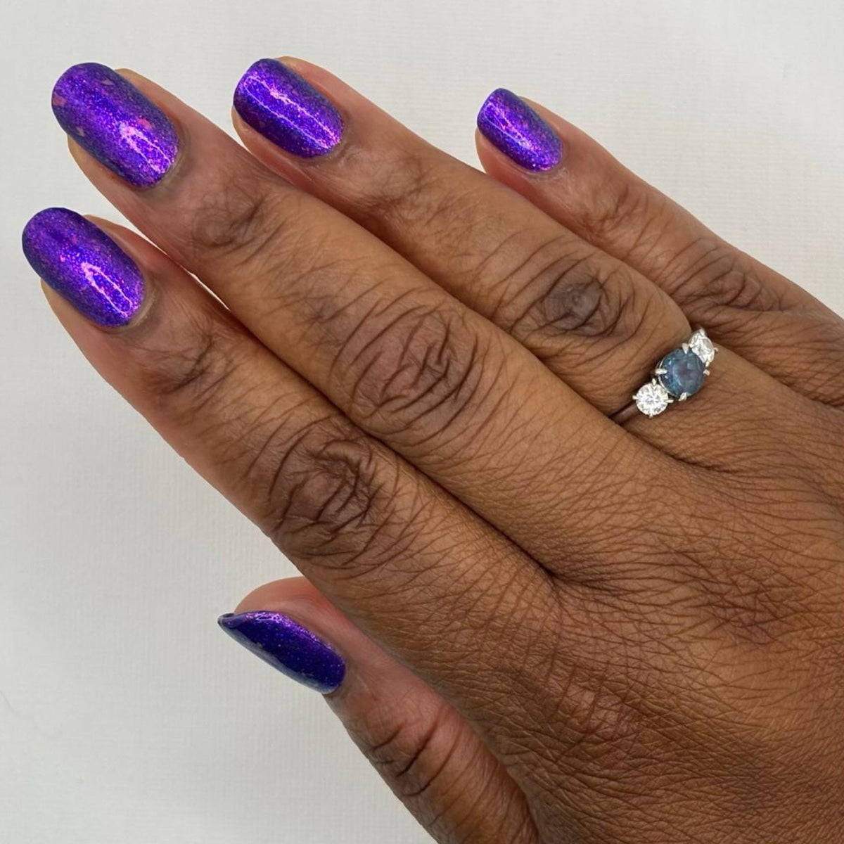 A close-up of a hand with dark skin with manicured nails painted in a shimmering metallic purple polish, wearing a silver ring with blue stones on one finger against a plain light background.