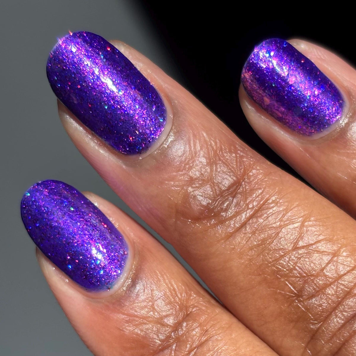 Close-up of a hand with dark skin tone manicured nails painted in a sparkling purple glitter nail polish.