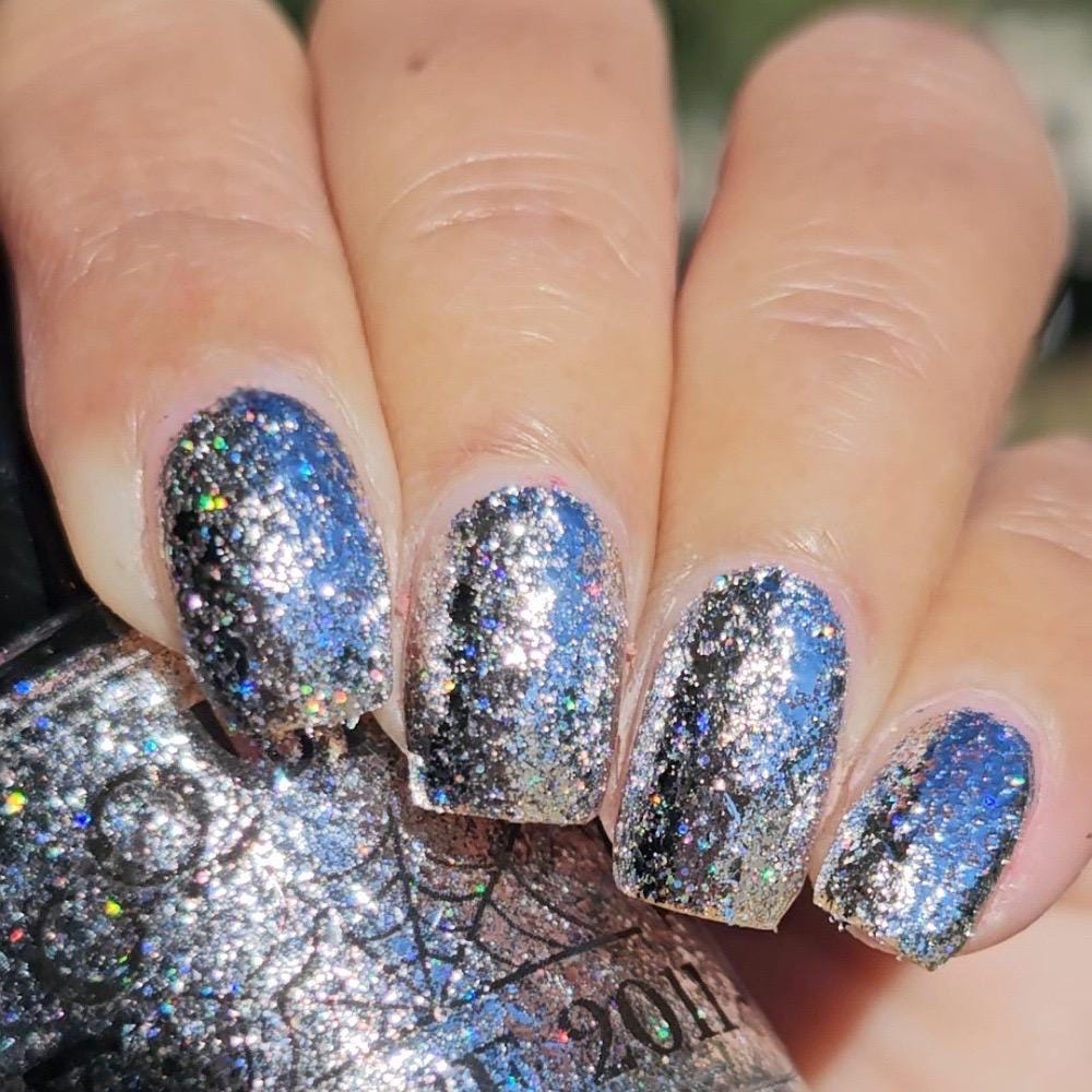 Close-up of a hand with manicured nails painted in a holographic silver glitter nail polish, with the matching glitter polish visible on a bottle in the foreground.