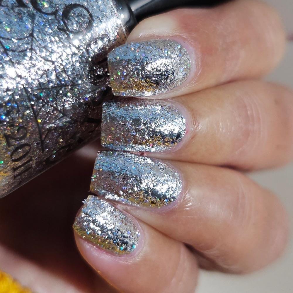 A close-up of a hand with nails painted in a sparkling silver glitter nail polish, holding a matching silver glitter nail polish bottle.