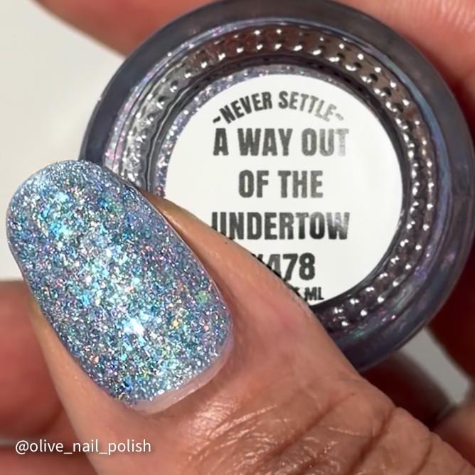 A close-up of a hand holding a bottle of nail polish, with the painted fingernail showing a glittery light blue and silver holographic finish. The pot label reads “Never Settle” and “A Way Out of the Undertow.