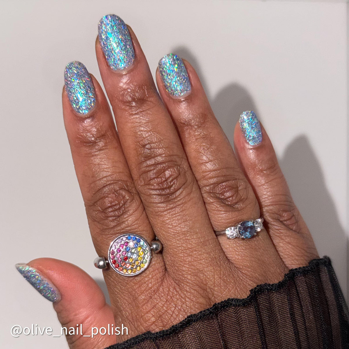 A close-up of a hand showcasing light blue holographic glitter nail polish, with silver rings on the fingers. The text '@olive_nail_polish' is visible in the bottom-left corner.