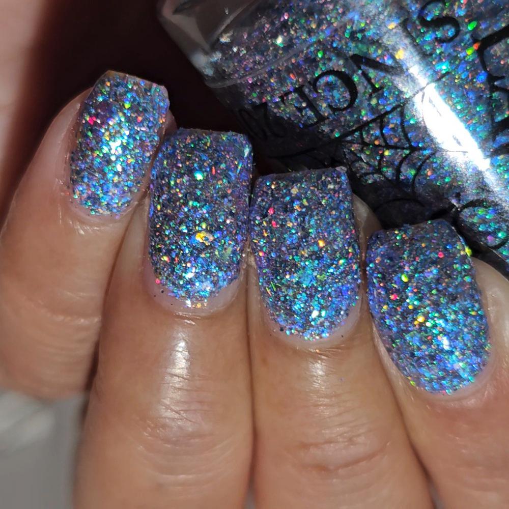 Close-up of a hand with manicured nails painted in a dense, iridescent holographic glitter nail polish, with the bottle partially visible in the background.