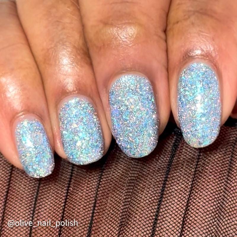Close-up of a hand with manicured nails painted in a sparkling light blue and white glitter nail polish, resting on a brown ribbed surface. A watermark in the corner reads '@olive_nail_polish'.