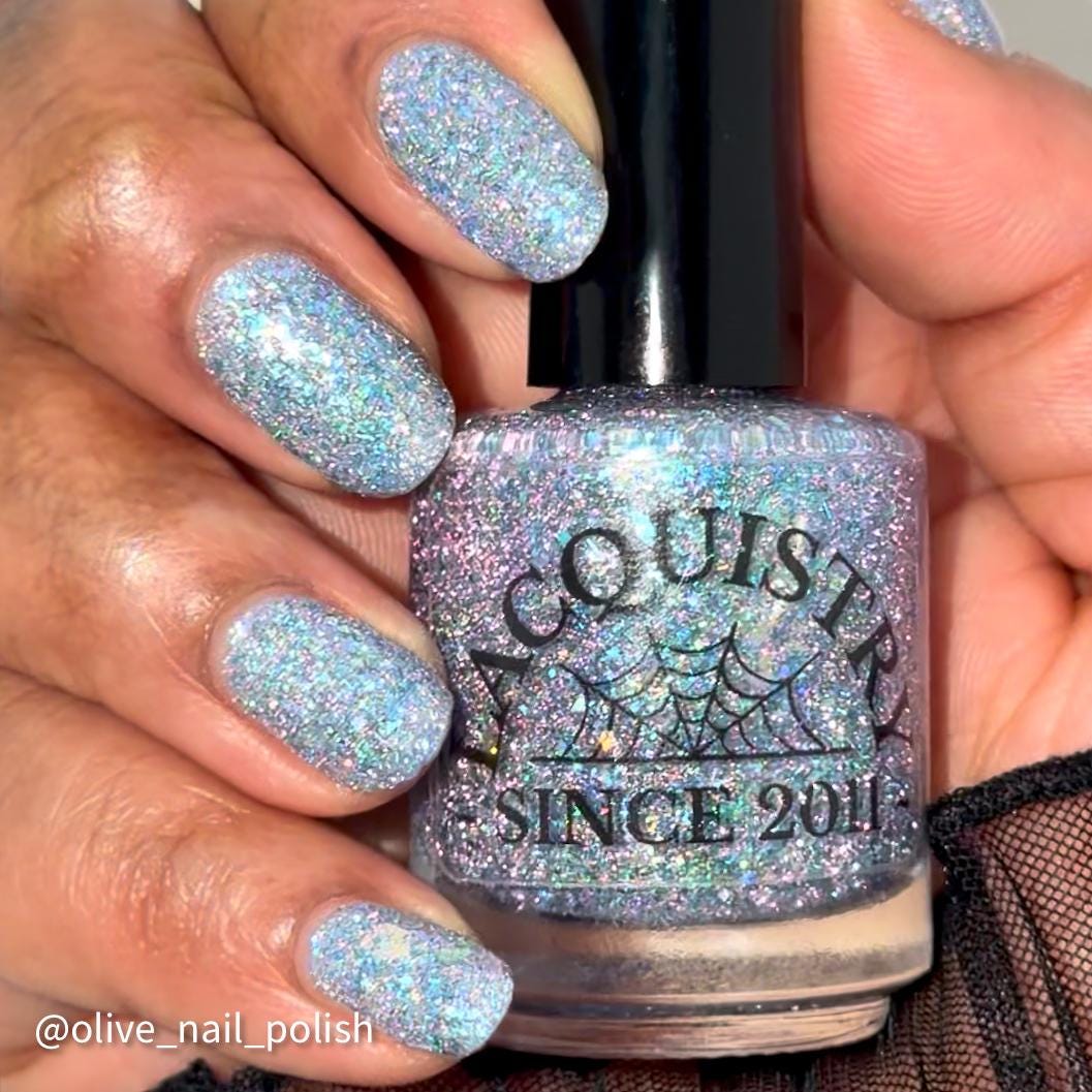 A close-up of a hand holding a bottle of nail polish. The nails are painted with a chunky, glittery light blue and silver polish, matching the holographic finish on the bottle. Bottle says Lacquistry Since 2011.