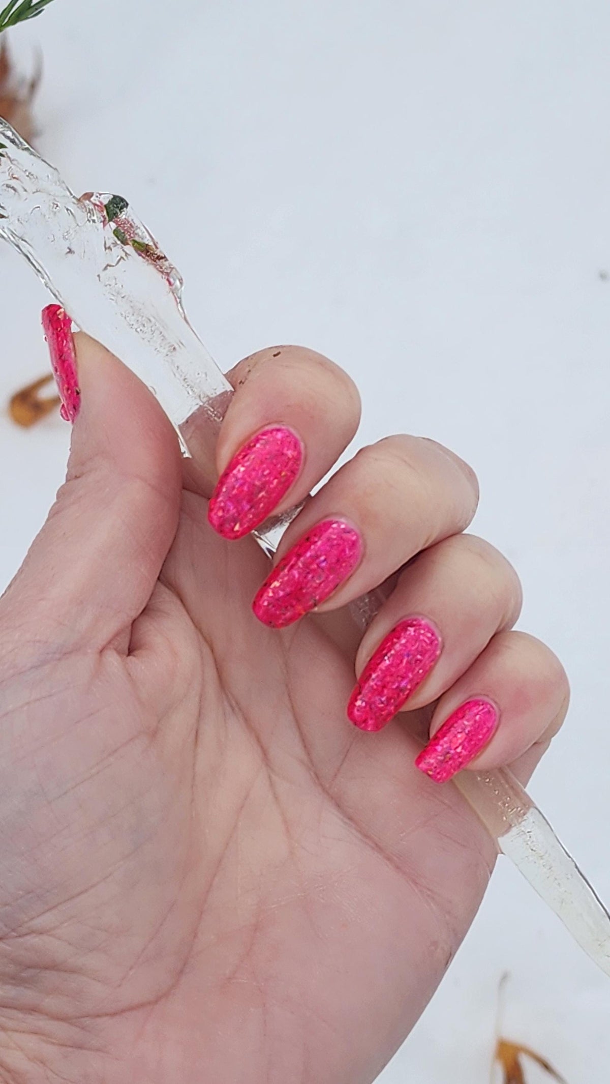 closeup of hand with light skin and long natural nails with neon pink flake polish holding an icicle against a white background
