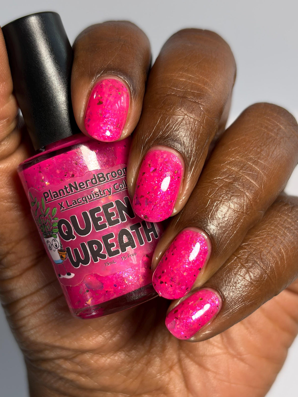 closeup of hand with dark skin and short nails with neon pink flake polish holding a nail polish bottle of the same color that says Queen Wreath PlantNerdBroom x Lacquistry