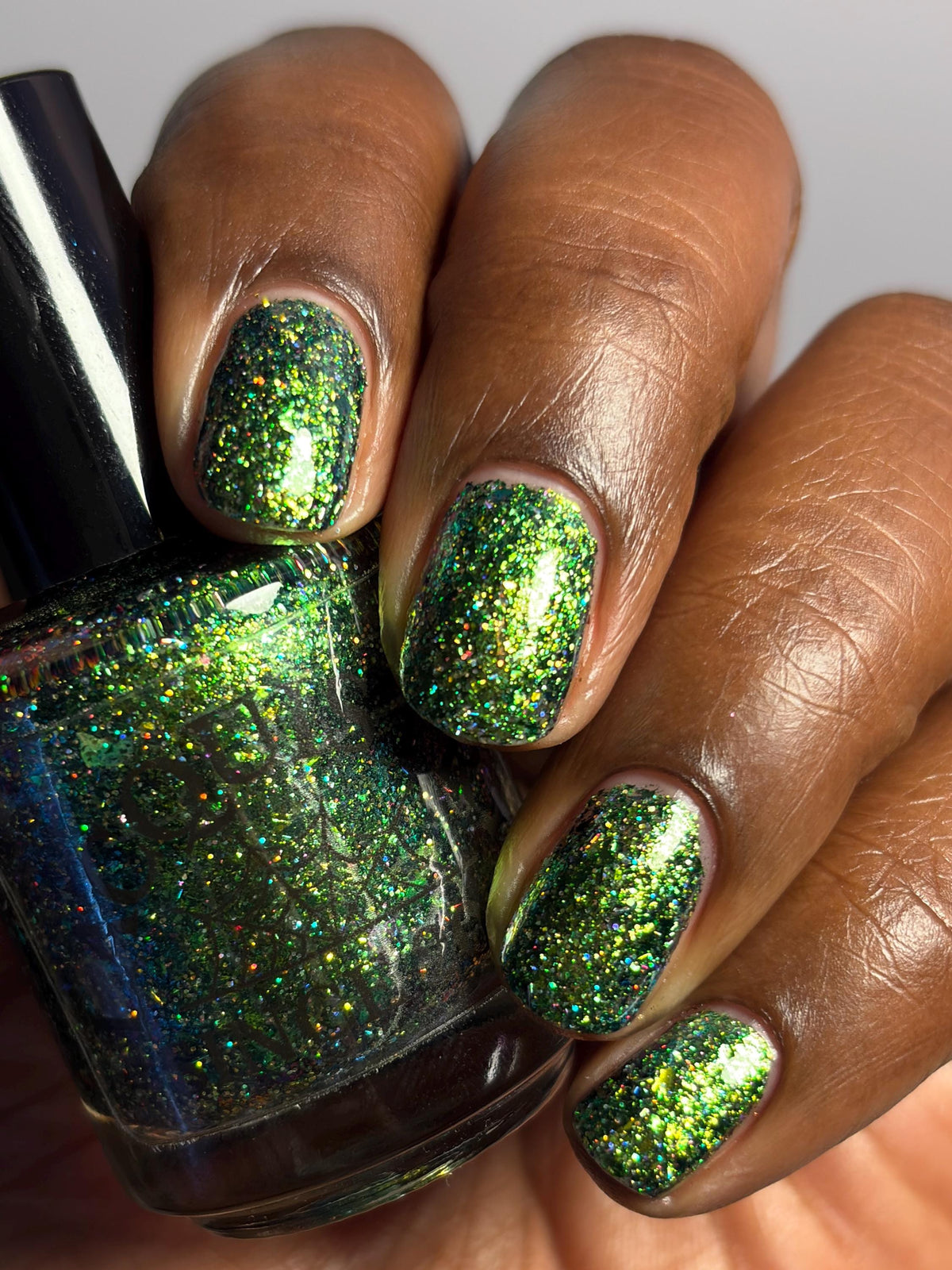 A close-up of a hand with nails painted in a sparkling green glitter nail polish, holding a matching green glitter nail polish bottle.