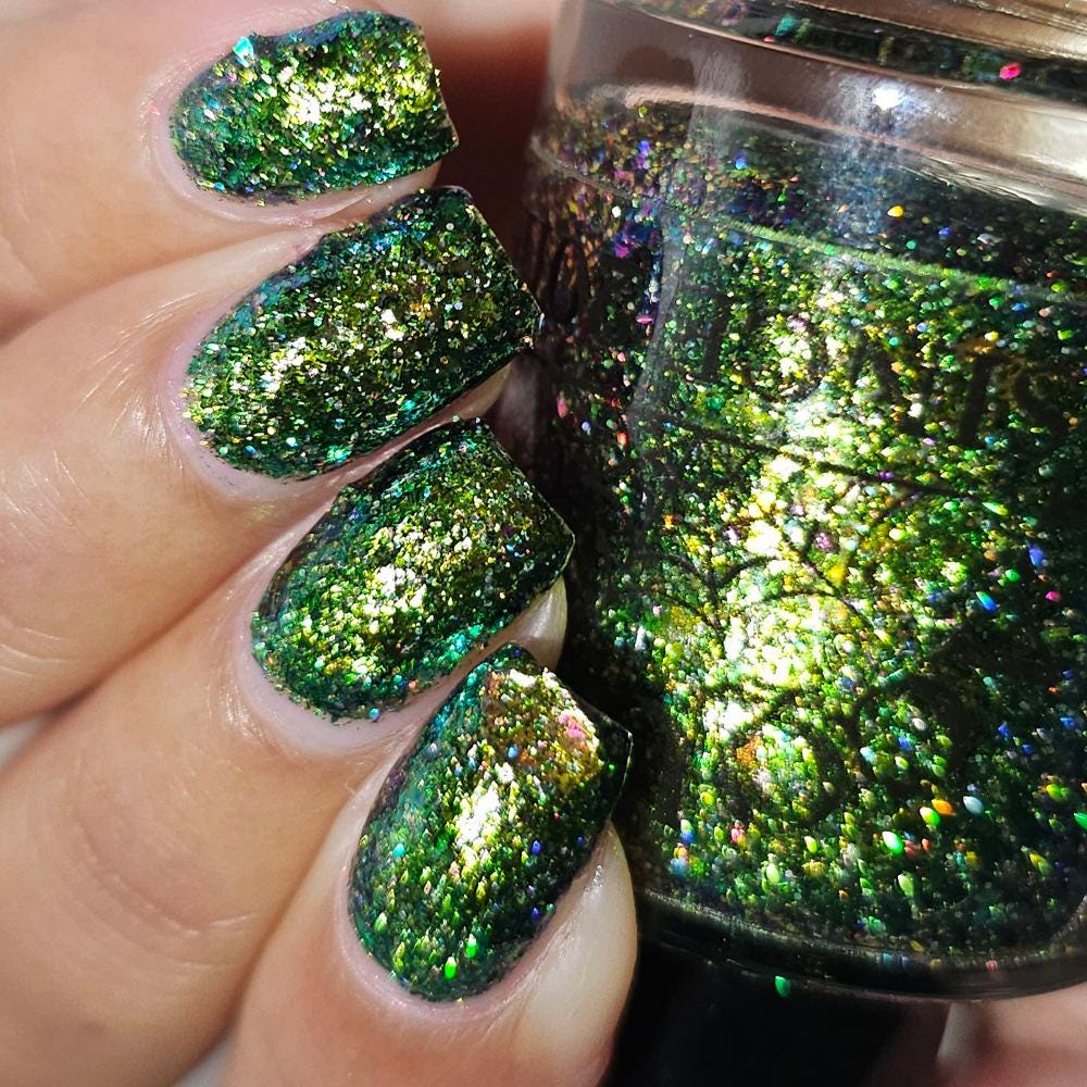 Close-up of manicured fingernails painted with a sparkling green holographic nail polish, with a matching bottle of the polish held beside the hand.