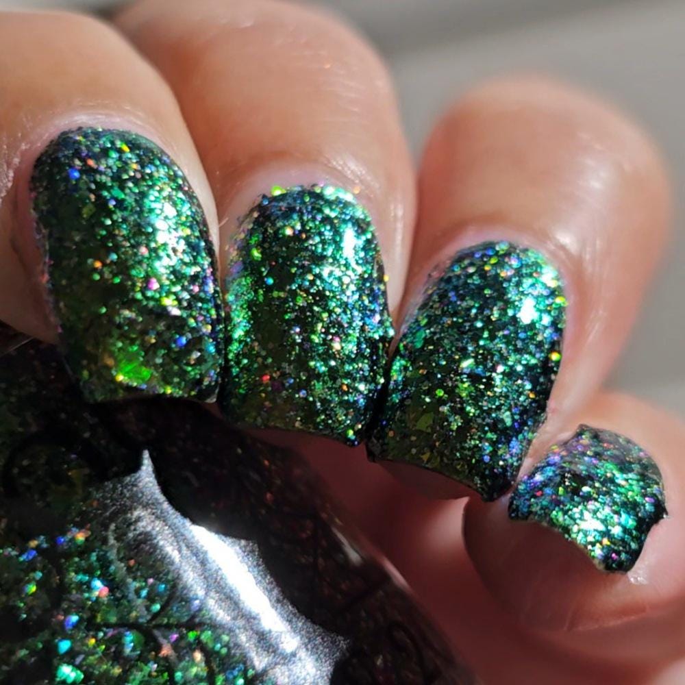 Close-up of a hand with manicured nails painted in a sparkling green holographic glitter nail polish, with the matching bottle partially visible in the foreground.