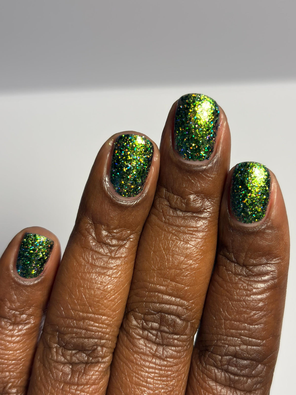 A close-up of a hand with medium to dark brown skin showcasing a vibrant green glitter nail polish on all fingernails, set against a neutral light gray background.