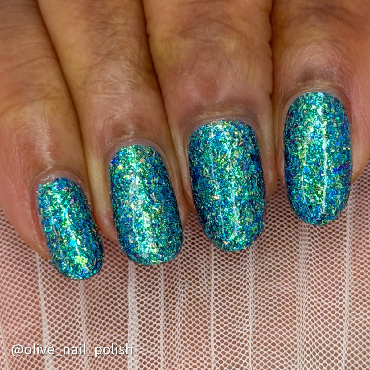 Close-up of a hand with almond-shaped nails painted in a dense, glittery holographic nail polish that shifts between teal, blue, and green. The hand is resting on a pink mesh surface, and a watermark in the bottom-left corner reads '@olive_nail_polish'.