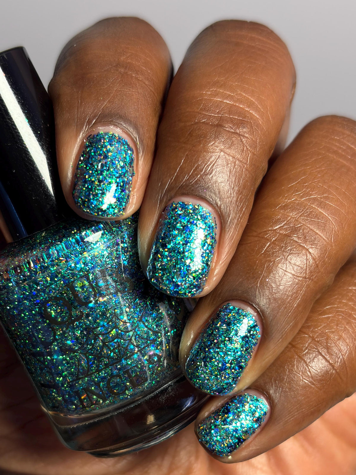 A close-up of a hand with medium-dark brown skin showcasing glittery teal blue nail polish, holding a matching glittery nail polish bottle.