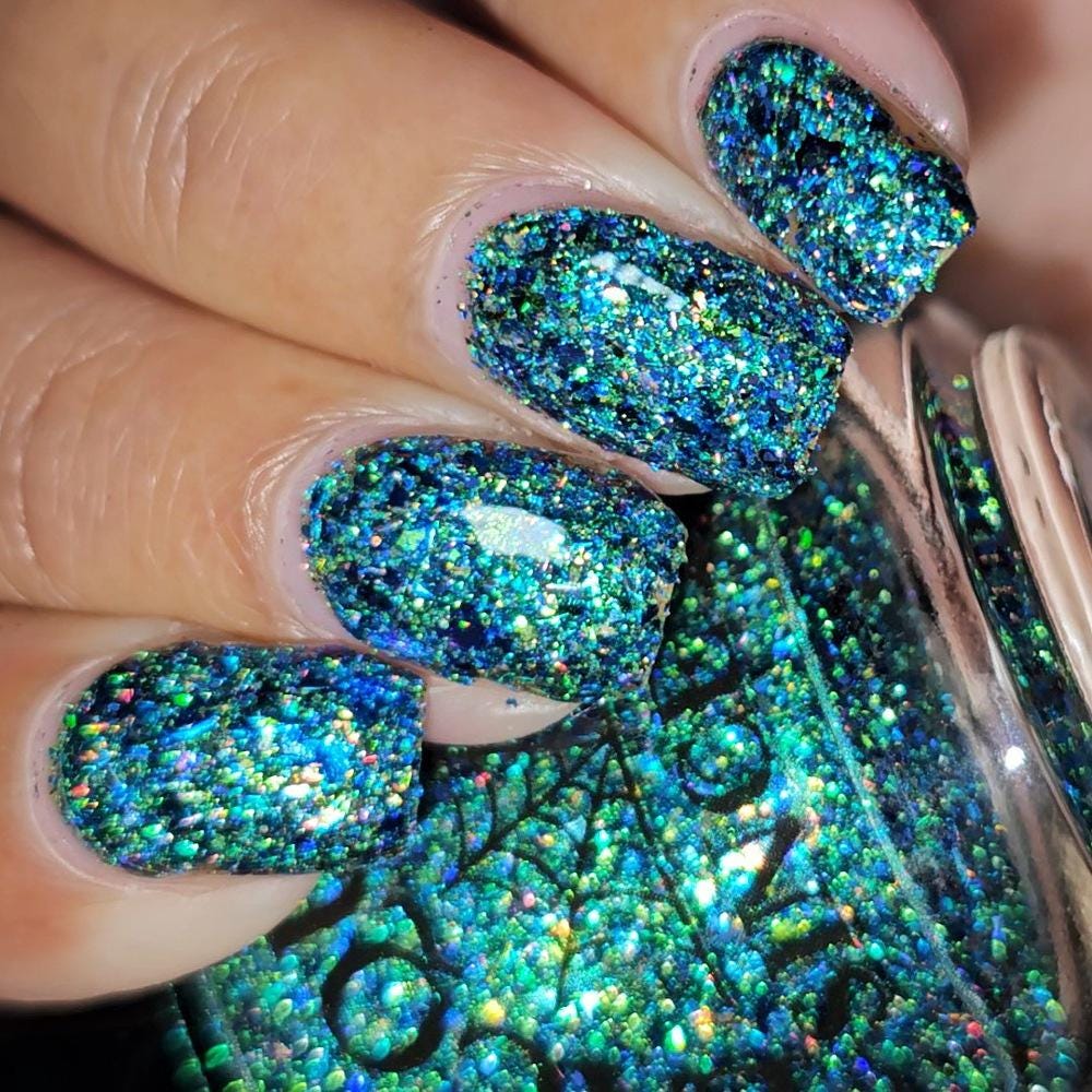 Close-up of a hand with manicured nails painted in a dense, glittery teal and blue holographic nail polish, with the matching polish bottle shown in the foreground.